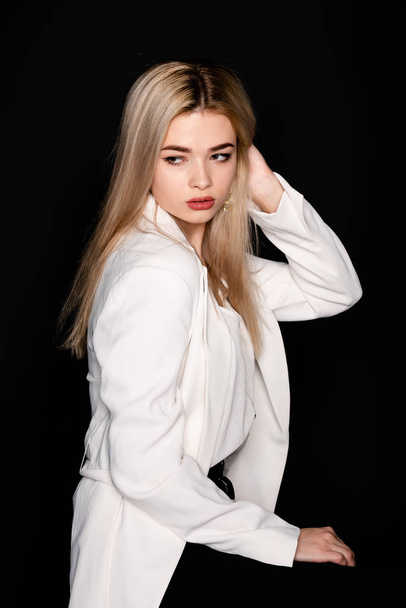 Young beautiful girl posing sitting on a chair in a white t-shirt and black trousers on a black background. Looks at the camera. - Zdjęcie, obraz