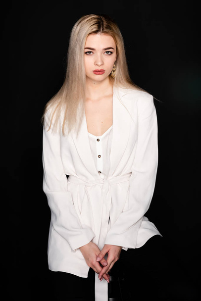 Young beautiful girl posing sitting on a chair in a white t-shirt and black trousers on a black background. Looks at the camera. - Foto, immagini