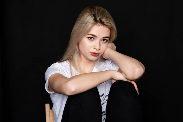 Young beautiful girl posing sitting on a chair in a white t-shirt and black trousers on a black background. Looks at the camera. - Фото, изображение