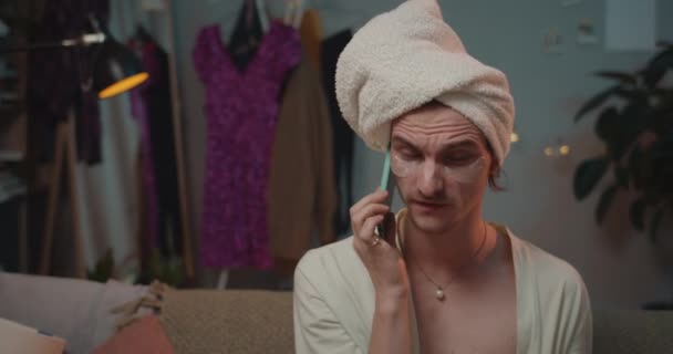 Close up of transgender man aggressively talking on smartphone and holding cigarette. Young trans man in patches and towel on head yelling on phone while sitting on sofa. - Imágenes, Vídeo