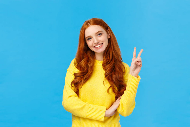 Waist-up shot cheerful tender and cute redhead modern woman in yellow sweater, tilt head and smiling lovely, showing peace sign, express positive emotions, joy and happiness, blue background - Foto, immagini