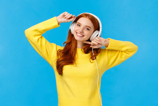 Технологии, интернет-магазины и концепция людей. Современная красивая рыжая женщина в желтом свитере, танцует беззаботно, слушает любимую певицу в наушниках, улыбается камера, синий фон
 - Фото, изображение