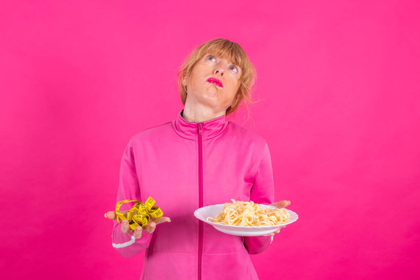 Frau mit Stressausdruck schaut auf Maßband und Teller mit Nudeln oder Hydraten mit Sportbekleidung auf rosa Hintergrund - Foto, Bild
