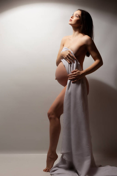 Naked pregnant woman with a white cloth on a gray background. - Foto, imagen