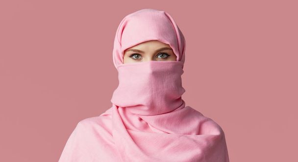 Portrait of young Muslim arabian woman wearing colorful hijab against pink background. Points finger to side. Space for text - Fotografie, Obrázek