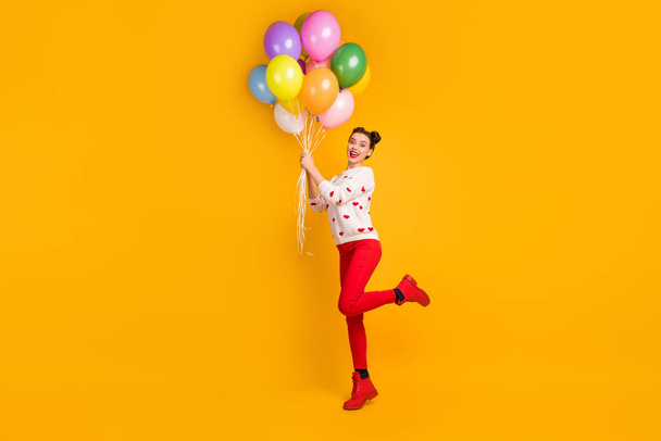 Im gonna fly. Full length profile photo of beautiful lady bring many colorful air balloons surprise party wear hearts pattern sweater red pants shoes isolated yellow color background - Zdjęcie, obraz