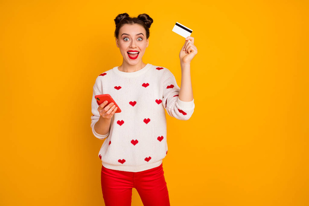Photo of pretty lady hold telephone plastic credit card making online purchase addicted shopper wear hearts pattern white pullover red pants isolated yellow color background - Fotoğraf, Görsel