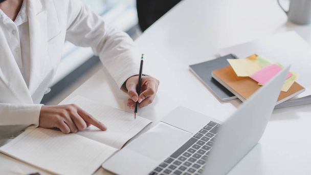 Plan recadré d'une femme d'affaires écrivant sur un carnet tout en travaillant sur un ordinateur portable avec des fournitures de bureau sur une table blanche dans une pièce de bureau
 - Photo, image