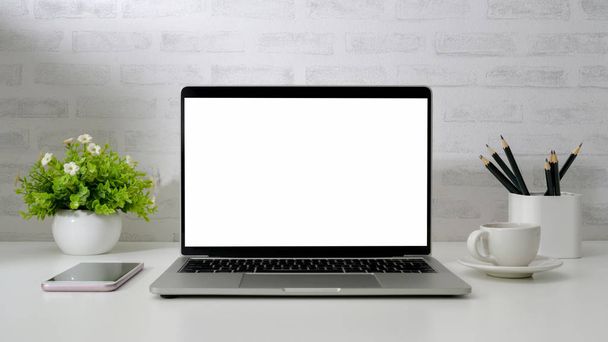 Close up widok obszaru roboczego z pustym ekranem laptop, smartfon, ołówki, filiżanka kawy i dzbanek na białym biurku z białej cegły tle ściany - Zdjęcie, obraz