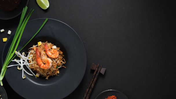Top view of Pad Thai, stir fly of Thai noodle with shrimp, egg, copy space and seasoning in black ceramic plate on black table  - Photo, Image