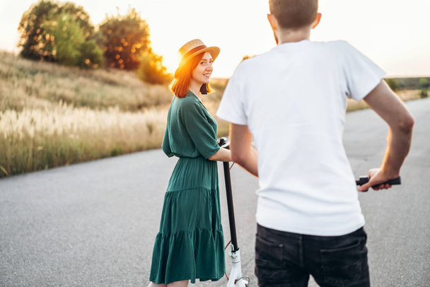 Happy couple walking on scooters. A young woman in dress and hat on sunset background. In the foreground, a man turns his back on the lens - Zdjęcie, obraz
