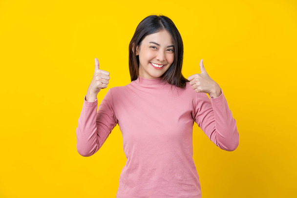 Asian smiling young woman doing Like action to be grad, banner or showing on isolated yellow color background, wearing winter cozy sweater indoors studio, happiness and fun, copy space concept - Photo, Image