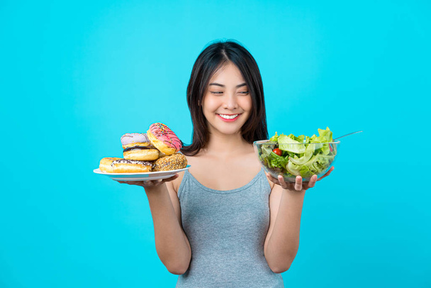 Atractiva mujer joven asiática sosteniendo y eligiendo entre el disco de rosquillas o ensalada de verduras en tazón vasos sobre fondo de color azul aislado, pérdida de peso y evitar la comida chatarra para la dieta y saludable
 - Foto, Imagen