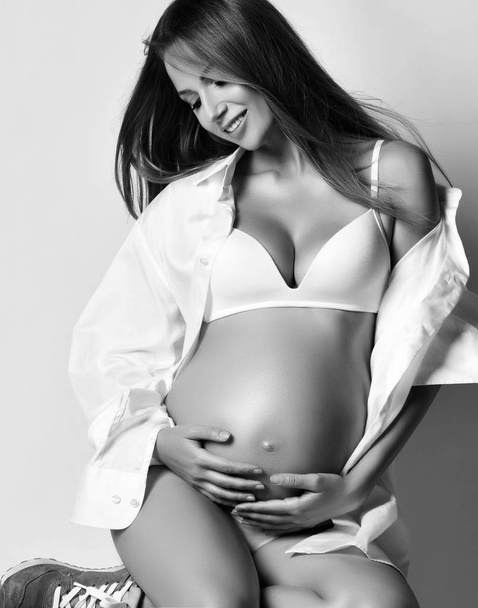 Young beautiful pregnant woman in white shirt and underwear and sneakers sitting, looking at belly and smiling - Фото, изображение