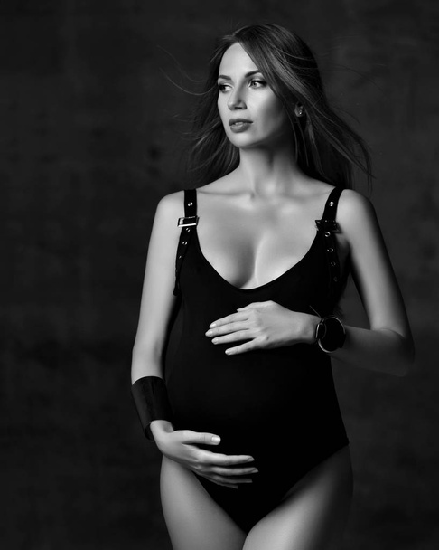 Young beautiful slim pregnant woman with long hair in black body and accessories standing, embracing belly in studio - Photo, Image