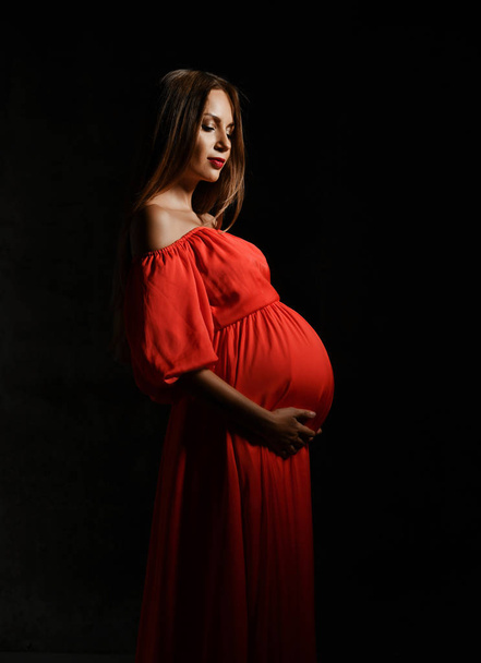 Young beautiful pregnant woman in red dress standing, embracing belly and smiling - Fotografie, Obrázek