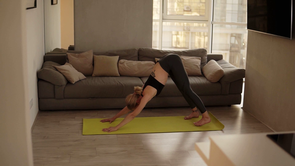 Mujer ejercitándose en casa practicando varias asanas de yoga, haciendo pose de perro boca abajo en salón sobre una esterilla de entrenamiento amarilla. Movimiento lento
 - Metraje, vídeo