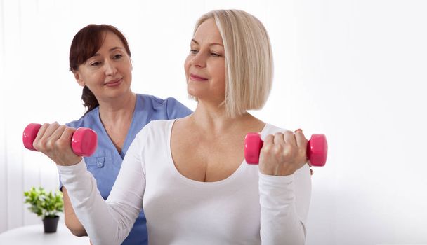 Picture of middle aged woman during rehabilitation in professional clinic. Rehabilitation, physiotherapy with dumbbells - Foto, Imagem