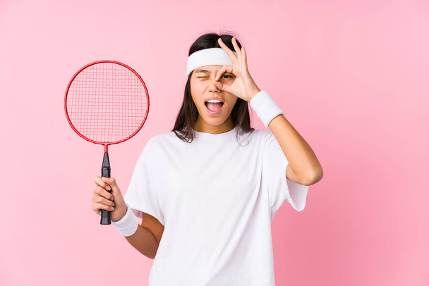 Jovem chinesa jogando badminton em um fundo rosa animado mantendo ok gesto no olho
. - Foto, Imagem
