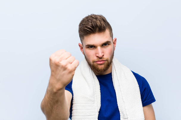 Mladý kavkazský sportovec s ručníkem ukazujícím pěst před kamerou, agresivní výraz obličeje. - Fotografie, Obrázek