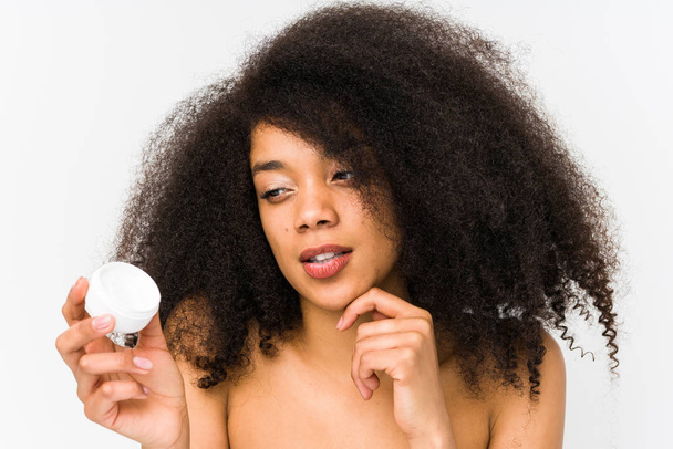 Jonge afro vrouw met een vochtinbrengende crème geïsoleerd kijken zijwaarts met twijfelachtige en sceptische uitdrukking. - Foto, afbeelding