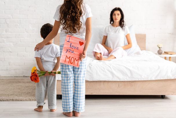 back view of brother and sister holding flowers and i love you mom card, and surprised woman sitting in bed - Photo, Image