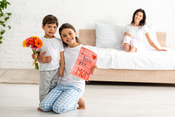 selective focus of children holding i love you mom card near mother sitting in bed - Valokuva, kuva