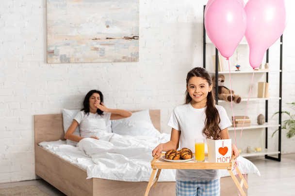cropped view of child holding tray with breakfast, mothers day card with heart sign and mom lettering, while mother stretching in bed - Foto, Bild