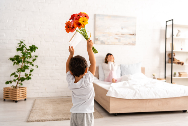 back view of boy holding flowers and mothers day card in raised hands, and mother sitting in bed and covering eyes with hands - Fotografie, Obrázek