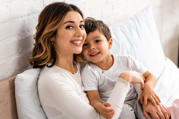 happy mother and son smiling at camera while sitting in bed in morning - Фото, изображение