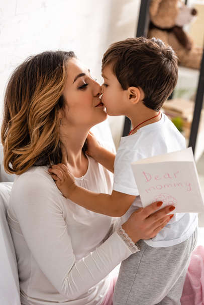 happy mother kissing adorable son while holding mothers day card with dear mommy inscription - Fotó, kép