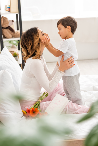 selective focus of cute boy touching face of happy mom sitting in bed near flowers and mothers day card - Photo, Image
