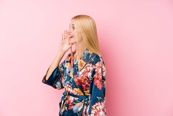 Mujer rubia joven con un pijama de kimono gritando y sosteniendo la palma cerca de la boca abierta
. - Foto, imagen