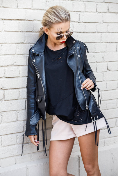 Beautiful blonde girl in black jacket and glasses posing on white brick wall background  - 写真・画像