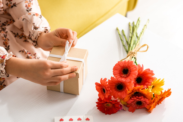 przycięty widok dziecka wiązania wstążki na dzień matki pudełko prezentów siedząc w pobliżu kwiatów na stole - Zdjęcie, obraz