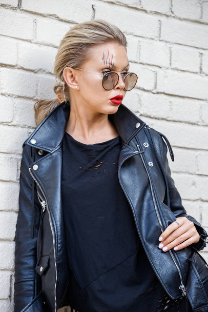 Beautiful blonde girl in black jacket and glasses posing on white brick wall background  - Фото, изображение