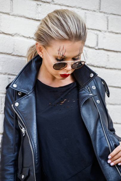 Beautiful blonde girl in black jacket and glasses posing on white brick wall background  - Fotografie, Obrázek
