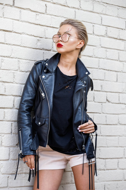Beautiful blonde girl in black jacket and glasses posing on white brick wall background  - Фото, изображение