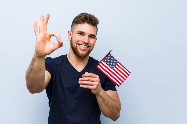 Jovem caucasiano segurando uma bandeira dos Estados Unidos alegre e confiante mostrando ok gesto
. - Foto, Imagem