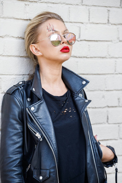Beautiful blonde girl in black jacket and glasses posing on white brick wall background  - Photo, image