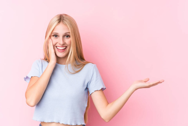 Young blonde woman on pink background holds copy space on a palm, keep hand over cheek. Amazed and delighted. - Photo, Image