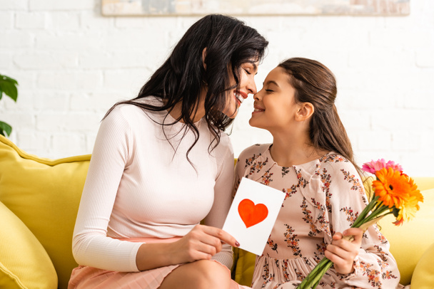 happy woman holding mothers day card with heart symbol, and daughter holding flowers while sitting on sofa and smiling face to face - Photo, Image