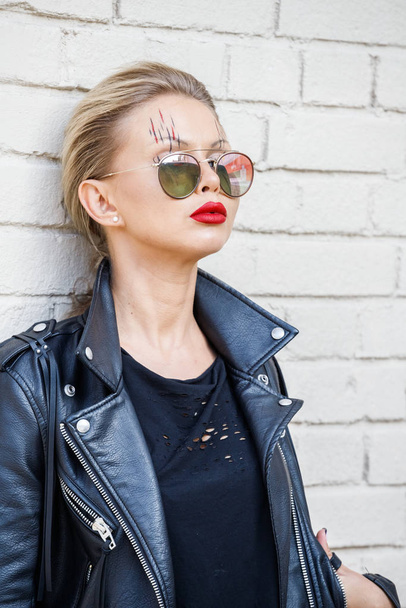 Beautiful blonde girl in black jacket and glasses posing on white brick wall background  - Foto, Bild