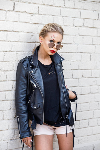 Beautiful blonde girl in black jacket and glasses posing on white brick wall background  - 写真・画像