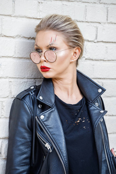 Beautiful blonde girl in black jacket and glasses posing on white brick wall background  - Photo, Image
