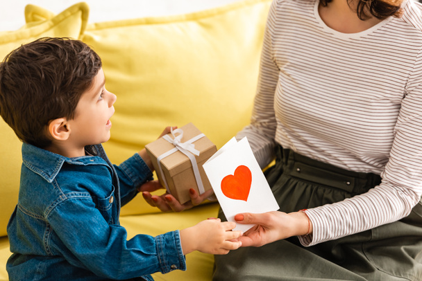 uroczy chłopiec prezentuje pudełko prezentów i kartka dzień matki z symbolem serca do mamy - Zdjęcie, obraz