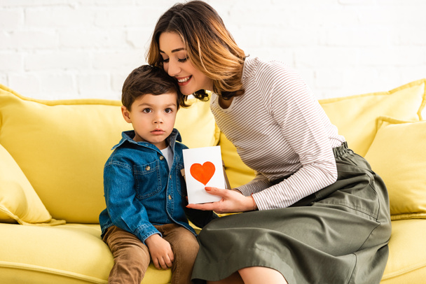femme gaie étreignant adorable fils tout en tenant la carte de jour des mères avec symbole de coeur
 - Photo, image