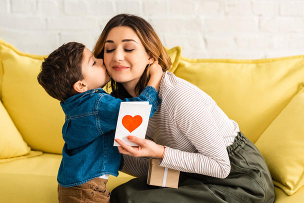 niedlicher Junge küsst glückliche Mutter mit Geschenkbox und Muttertagskarte mit Herzsymbol - Foto, Bild
