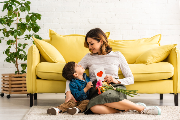 smiling woman looking at adorable son while sitting on floor with mothers day card and flowers - Foto, Imagen