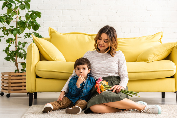 happy woman sitting on floor with flowers and hugging adorable son on mothers day - Foto, Bild